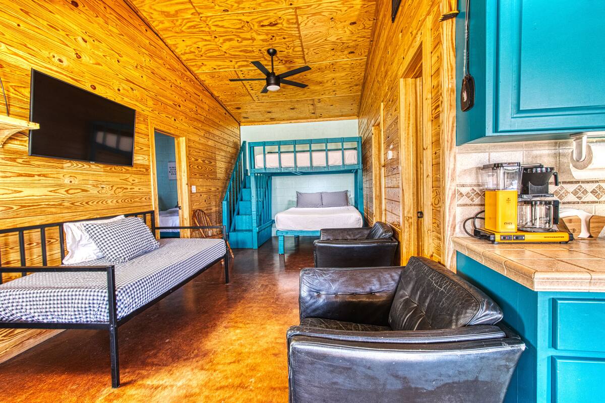 A cabin interior with a bunk bed, a sofa, a dining area with a coffee machine, and a television mounted on the wall.