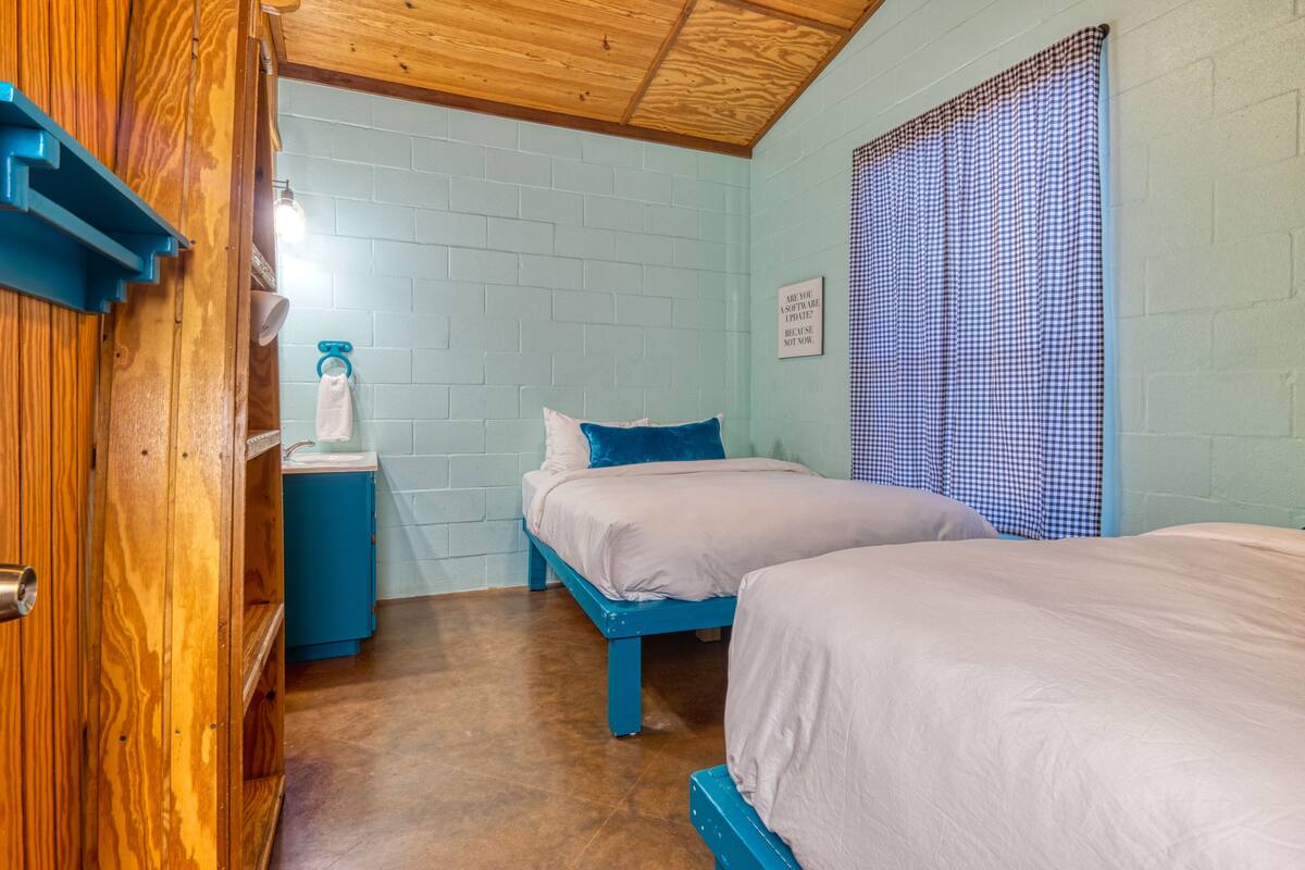 A bedroom with two beds, a wooden door, a blue bench, a white wall, and a checkered curtain.