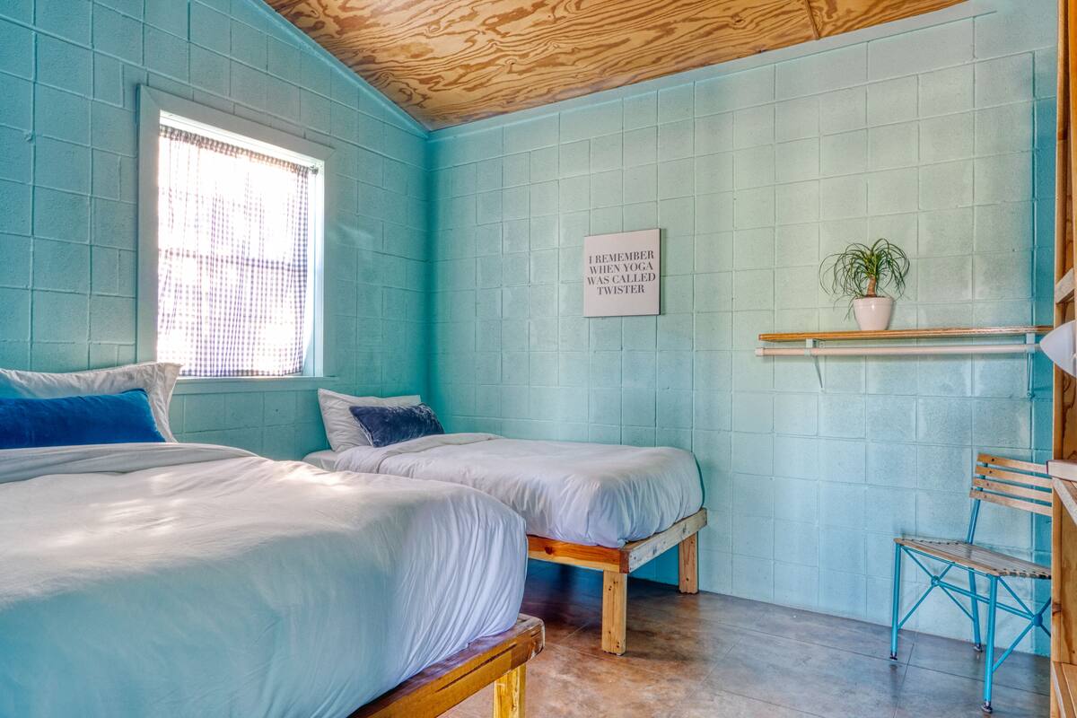 A room with two beds, a window with blinds, a wall-mounted sign, a potted plant, and a wooden chair.