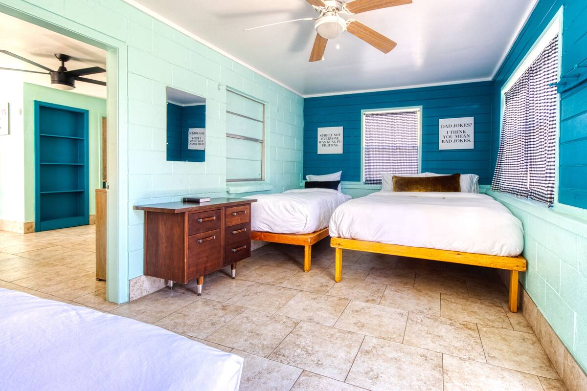A bedroom with two beds, a wooden dresser, a ceiling fan, and a window with blinds.