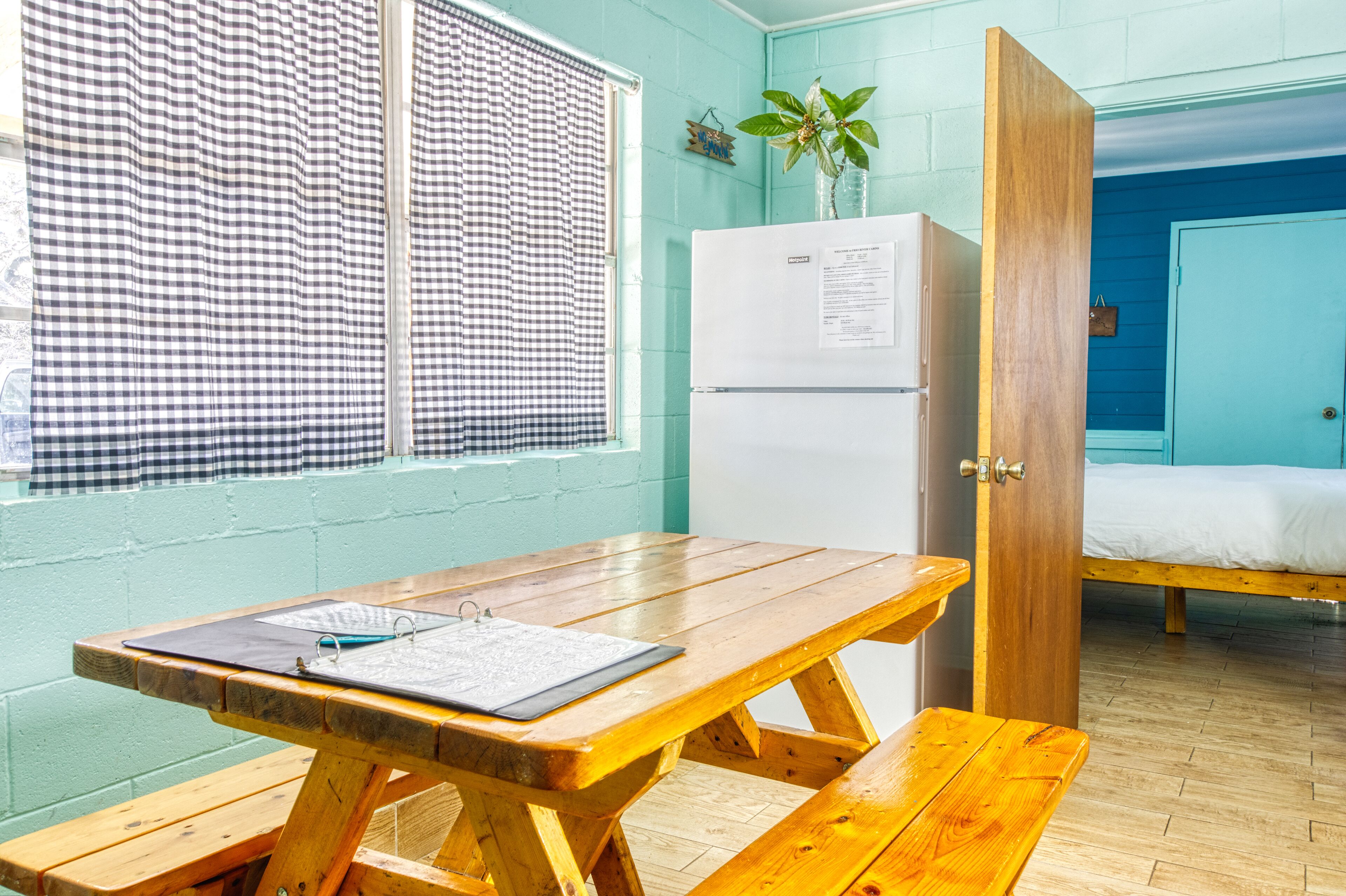 A room with a wooden table, a refrigerator, a bed, and a window with checkered curtains.