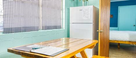 A room with a wooden table, a refrigerator, a bed, and a window with checkered curtains.
