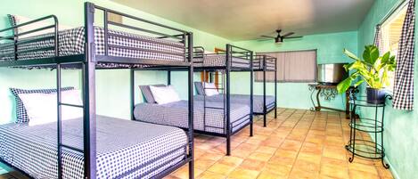 Une chambre de dortoir avec des lits superposés, un ventilateur de plafond, une fenêtre avec des stores et une plante en pot.