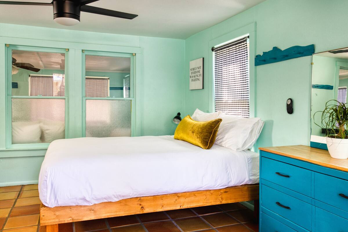 A bedroom with a bed, a wooden headboard, a blue dresser, a ceiling fan, and a window with blinds.