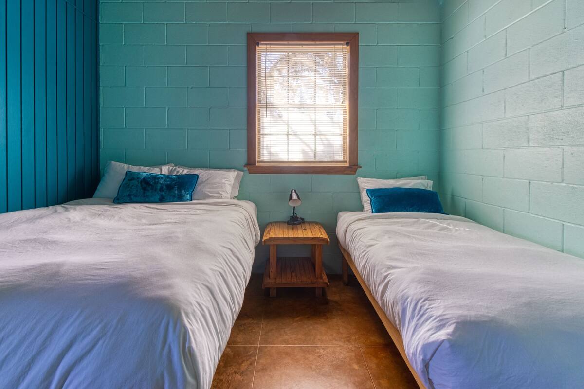 A room with two beds, a window with blinds, and a small wooden table.