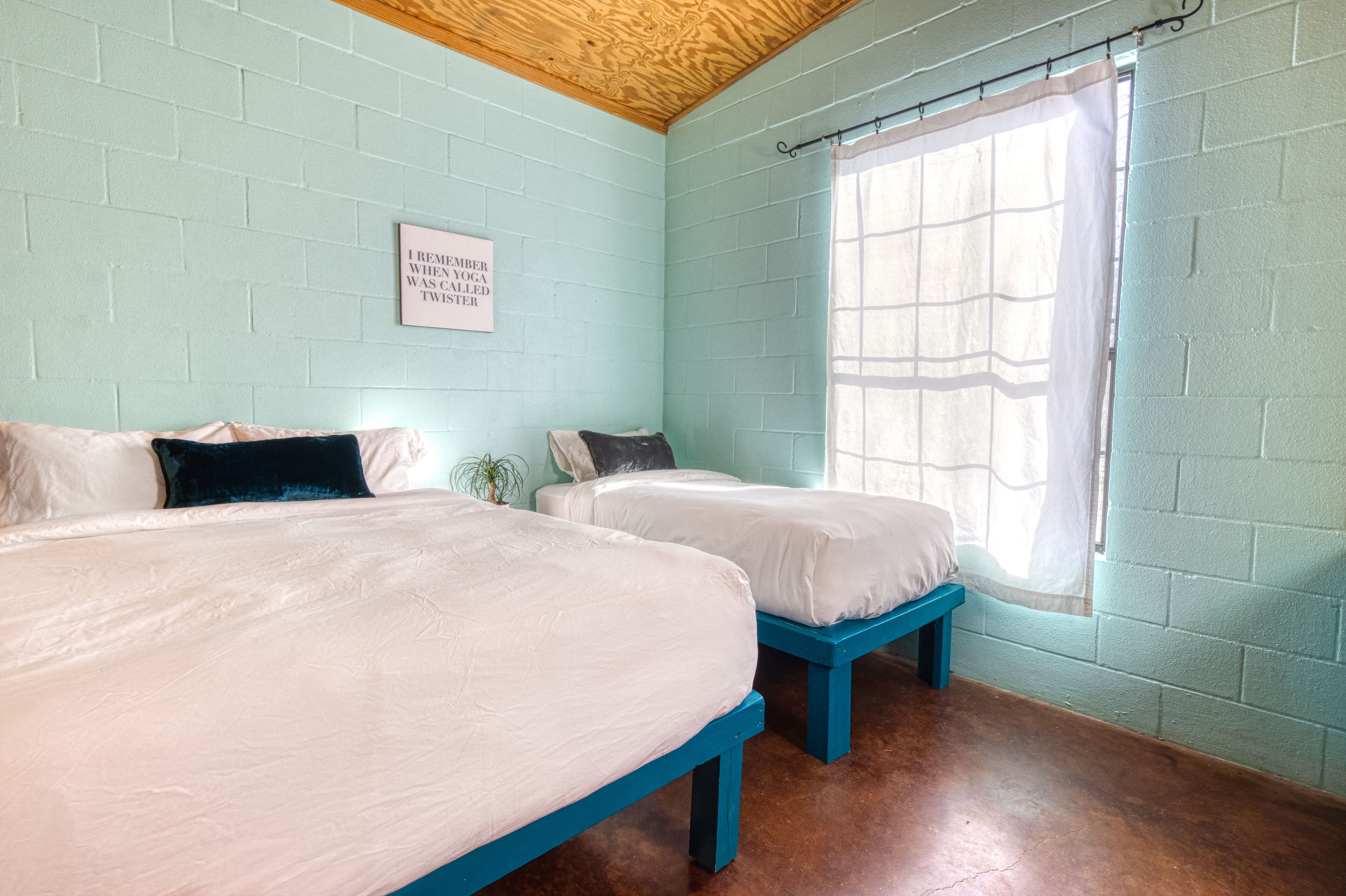 A room with two beds, a wooden ceiling, light blue brick walls, and a window with a sheer curtain.