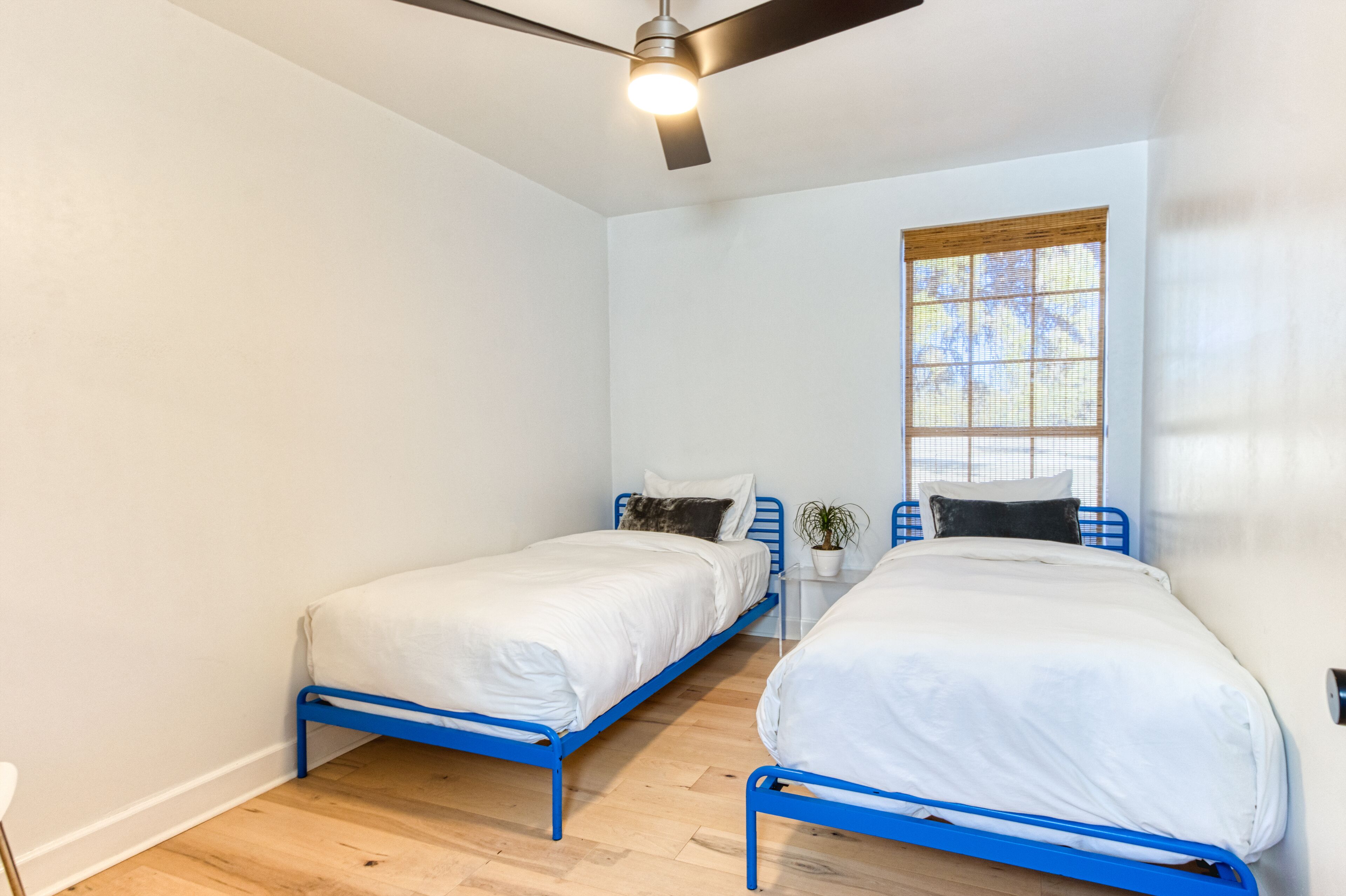 Un dormitorio con dos camas, un ventilador de techo, una ventana con persianas y una mesita con una planta.