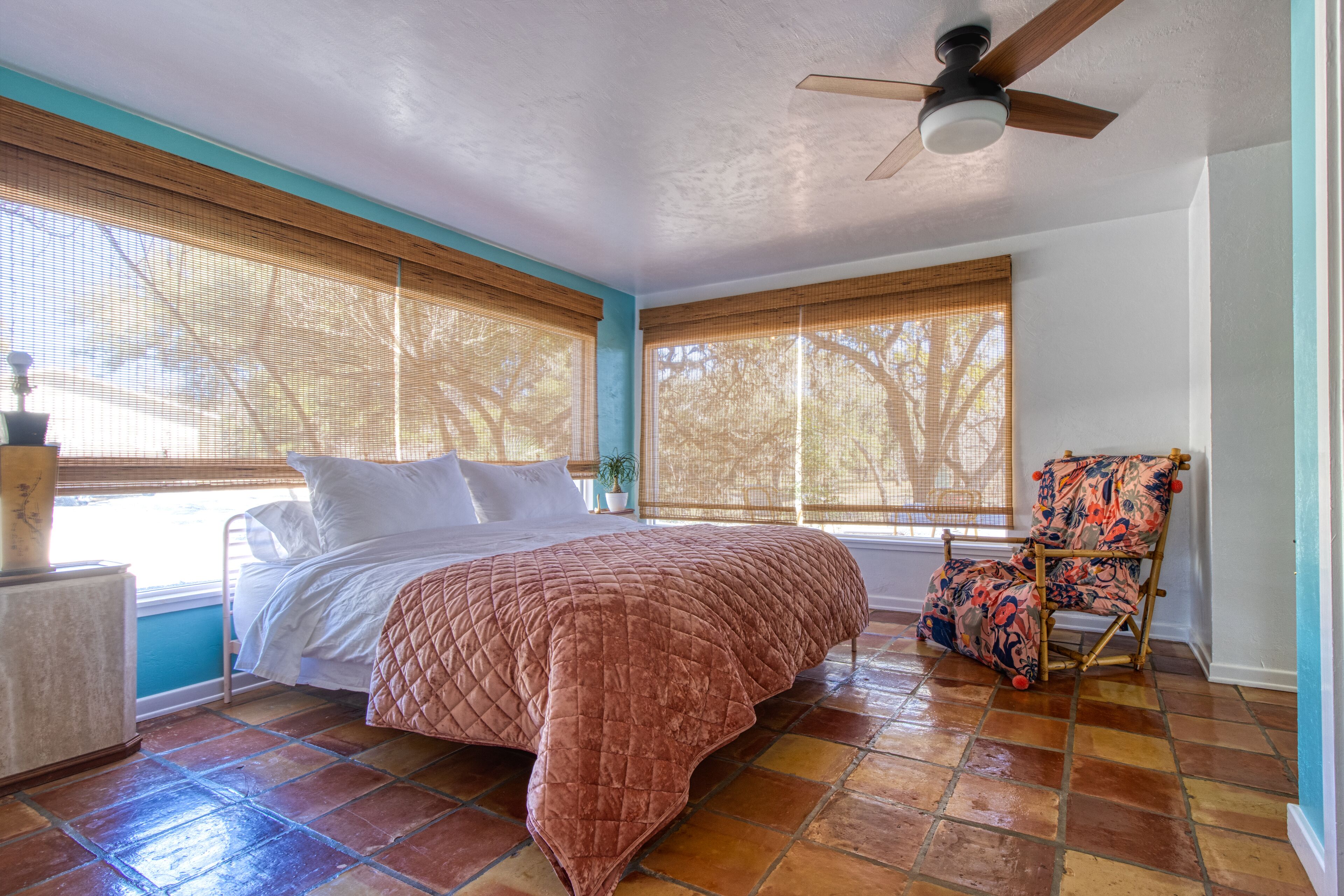 A bedroom with a bed, a chair, a ceiling fan, and large windows with blinds.