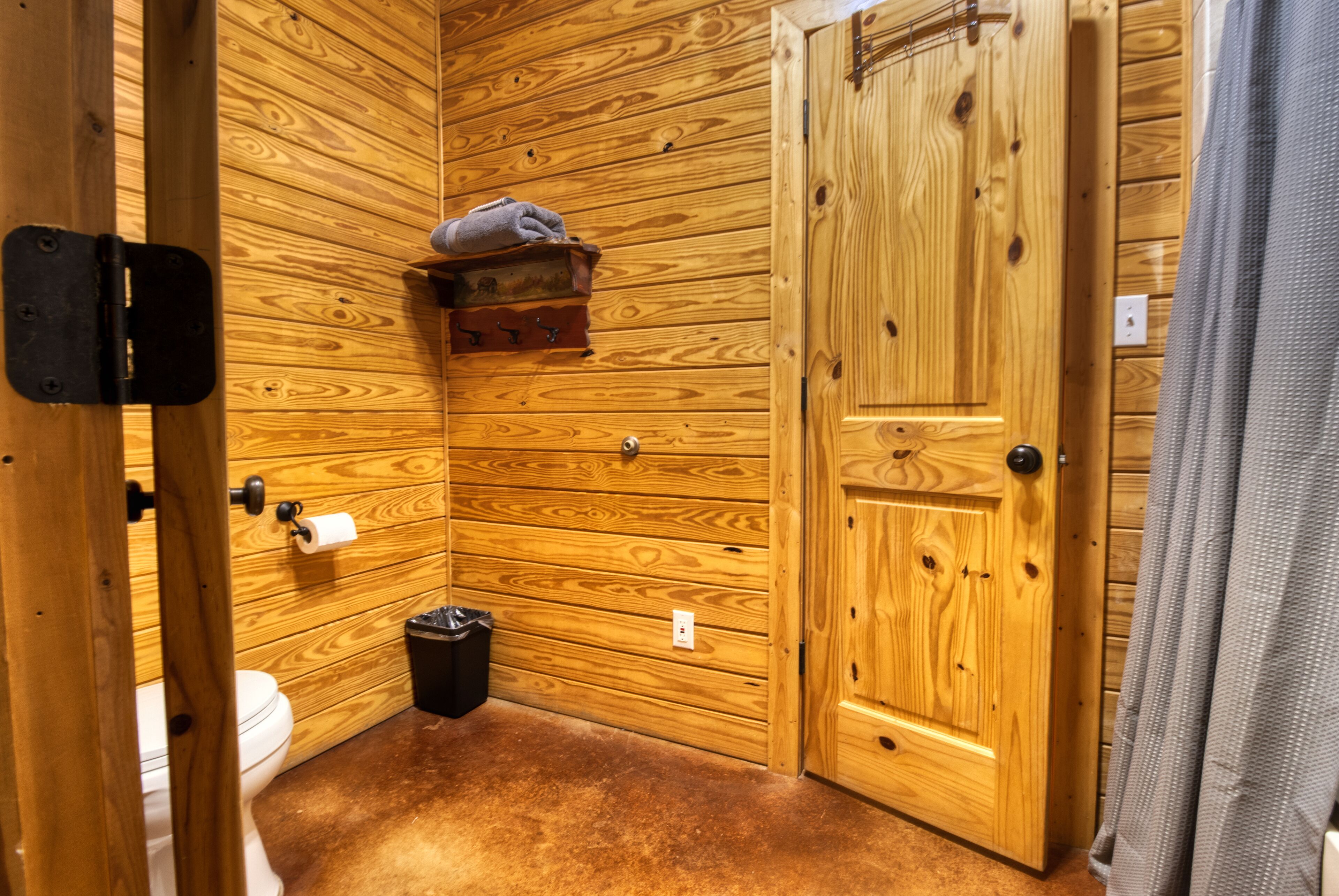 A wooden-paneled bathroom with a toilet, a trash bin, and a wooden shelf with a towel.