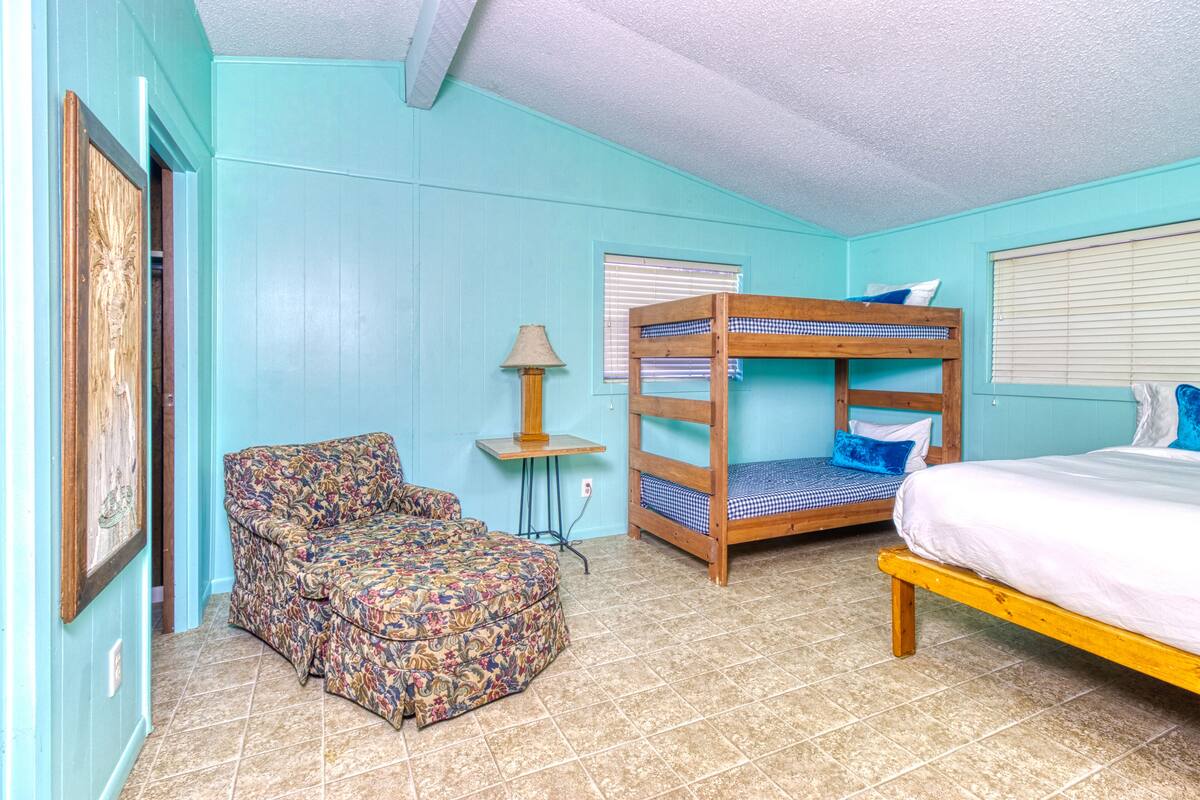 A room with a bunk bed, a patterned chair, a small table with a lamp, and a bed with white bedding.