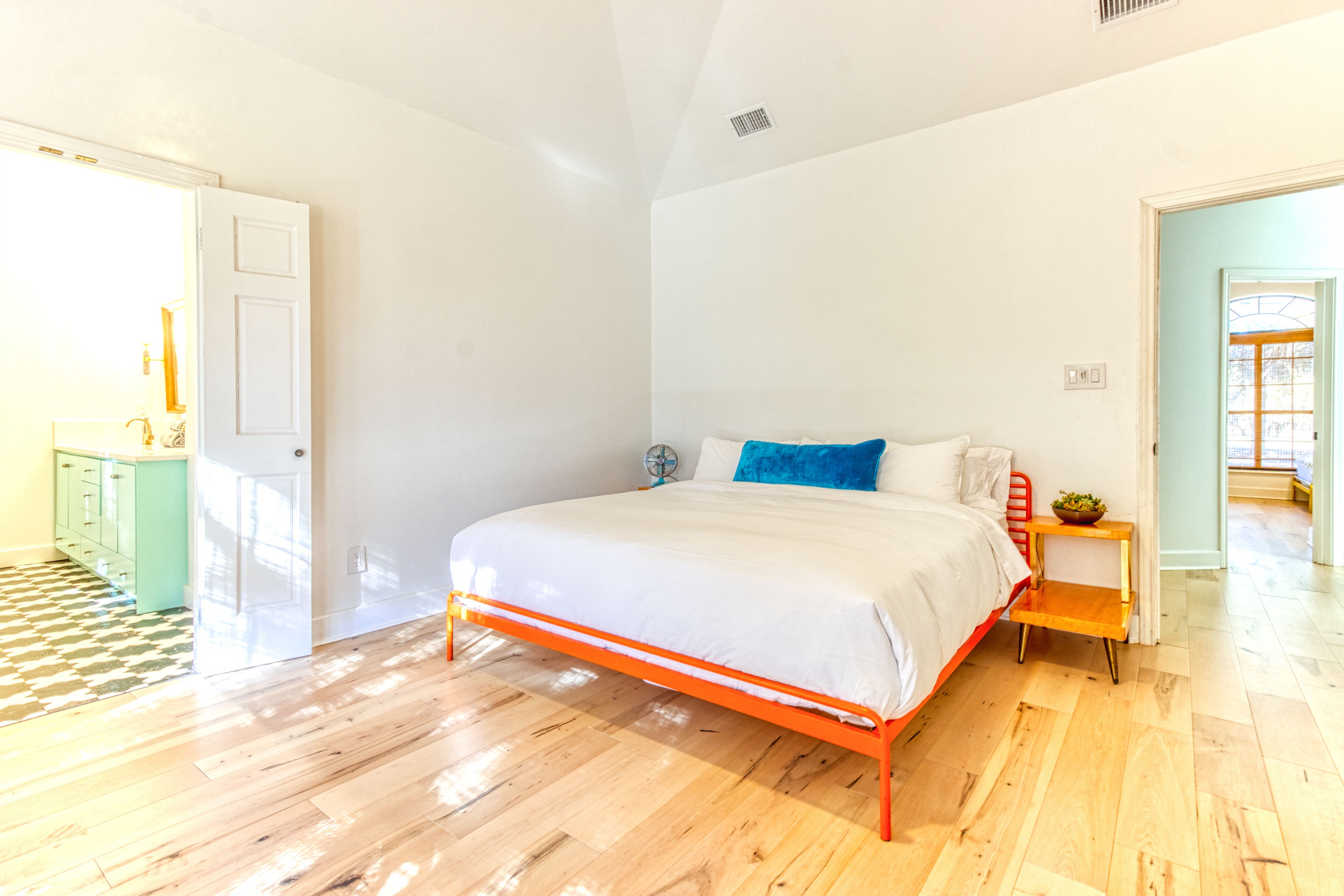 Une chambre avec un plancher en bois, un lit blanc avec un cadre en métal orange, une petite table de chevet avec une plante, et une vue sur un couloir.