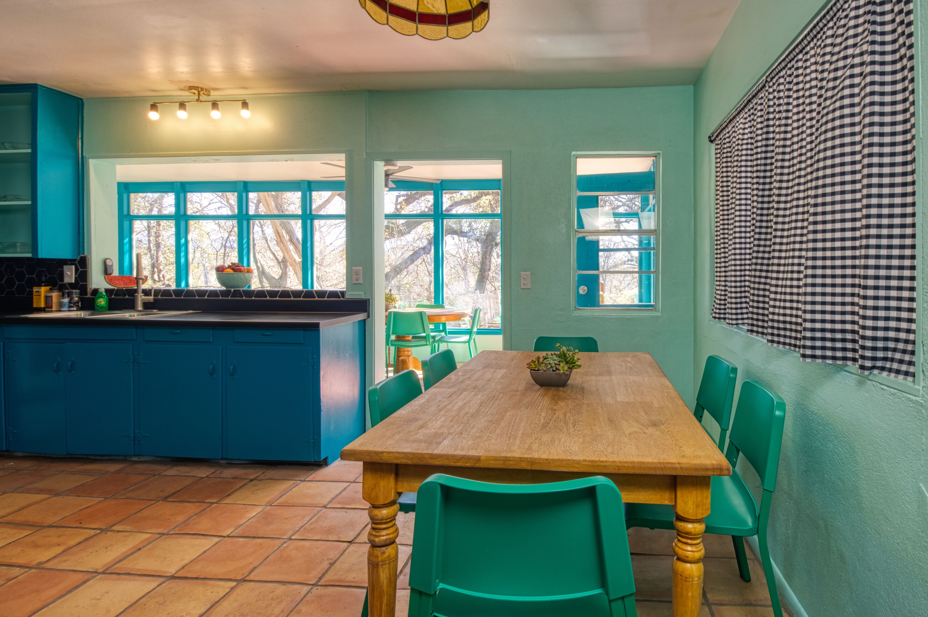 Un comedor con una mesa de madera y sillas verdes, una cocina con armarios azules, y un ventanal con persiana a cuadros.