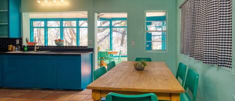 Un comedor con una mesa de madera y sillas verdes, una cocina con armarios azules, y un ventanal con persiana a cuadros.