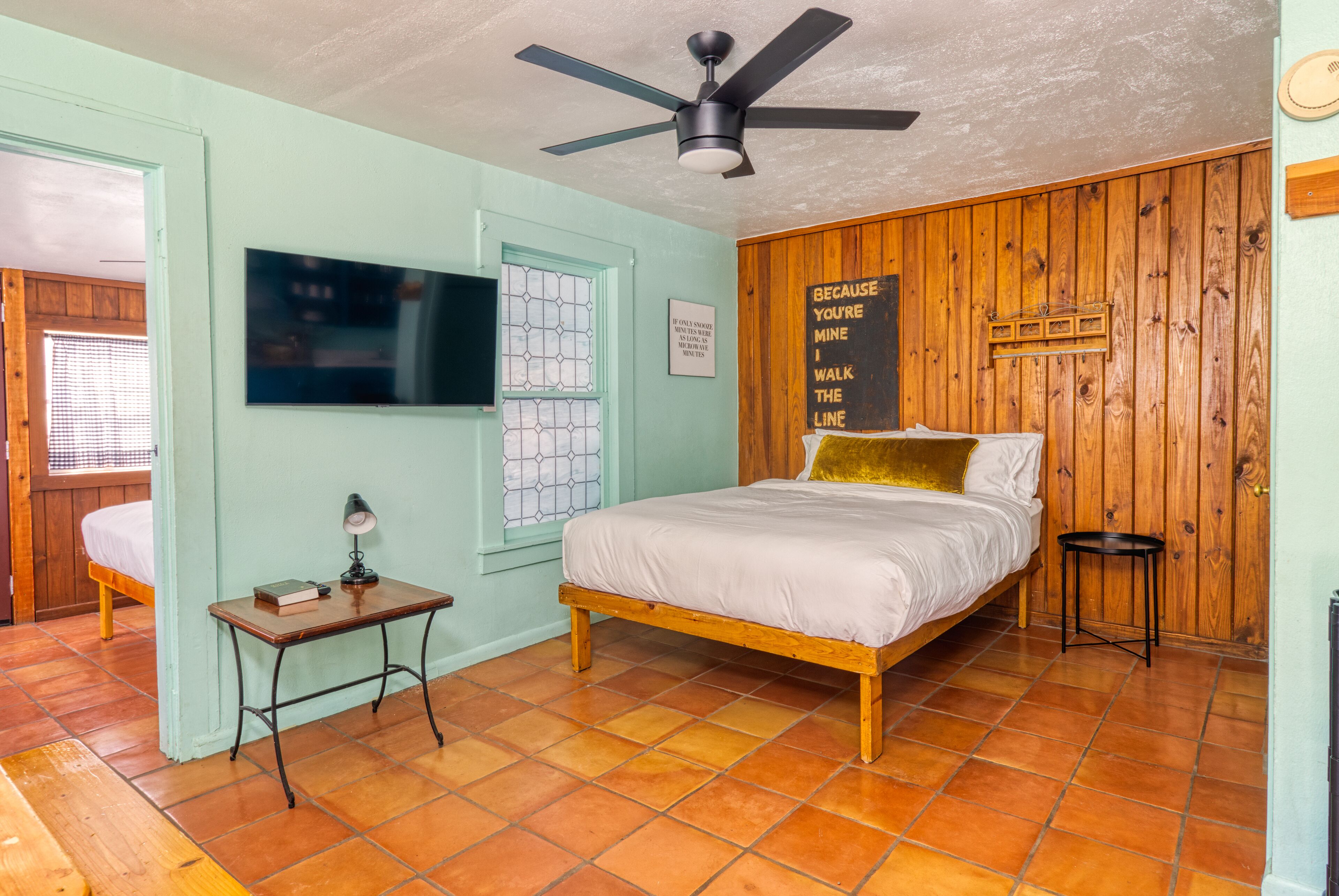 A bedroom with a bed, a ceiling fan, a TV, a small table, and a wooden wall with a sign.