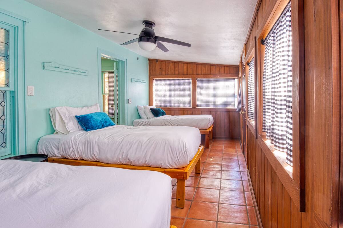 A room with three single beds, wooden flooring, a ceiling fan, and large windows with blinds.