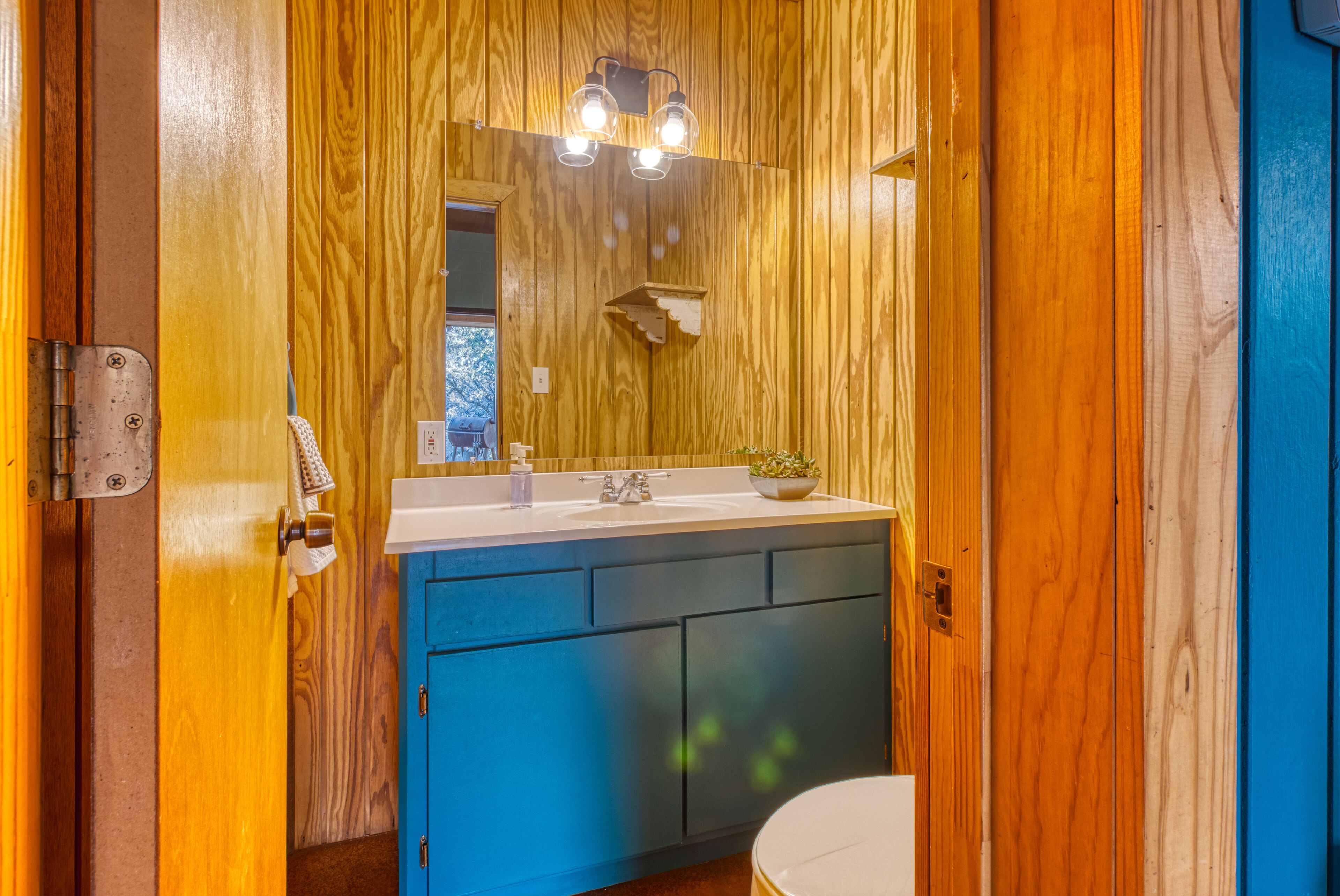 A bathroom with wooden walls, a blue cabinet, a white sink, and a mirror with lights above it.