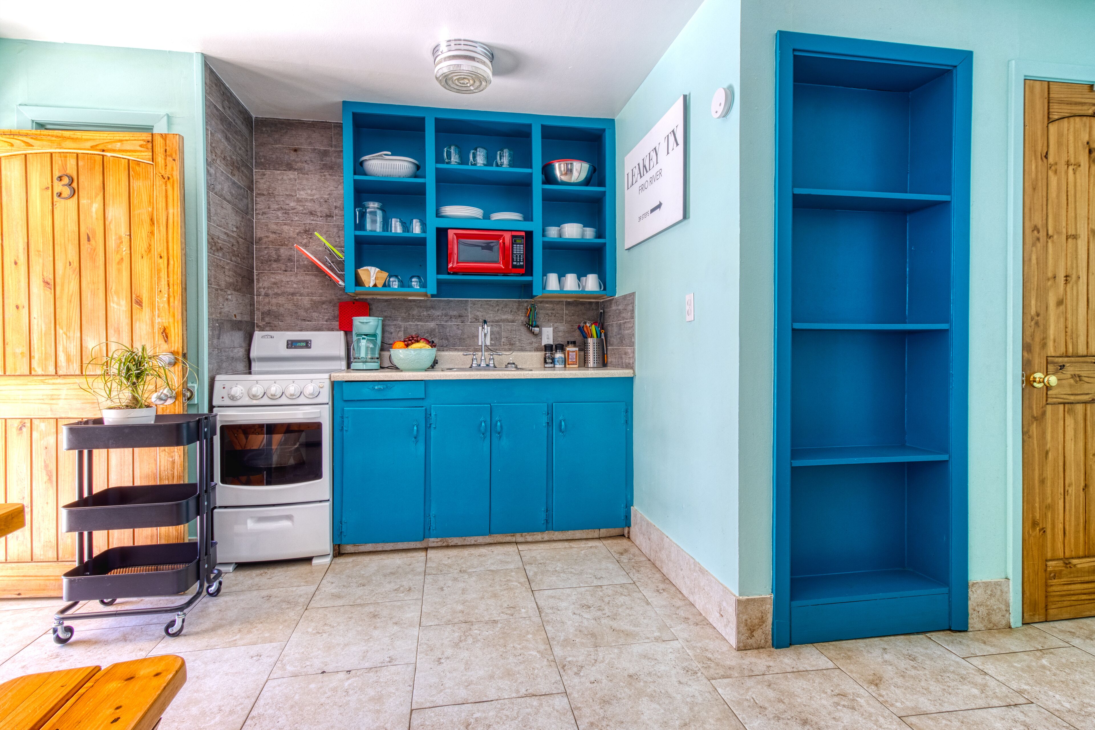 A kitchen with blue cabinets, a microwave, and a sink. There is a wooden door with a number 3 sign, a cart with trays, and a sign that reads "LEAKEY TX".