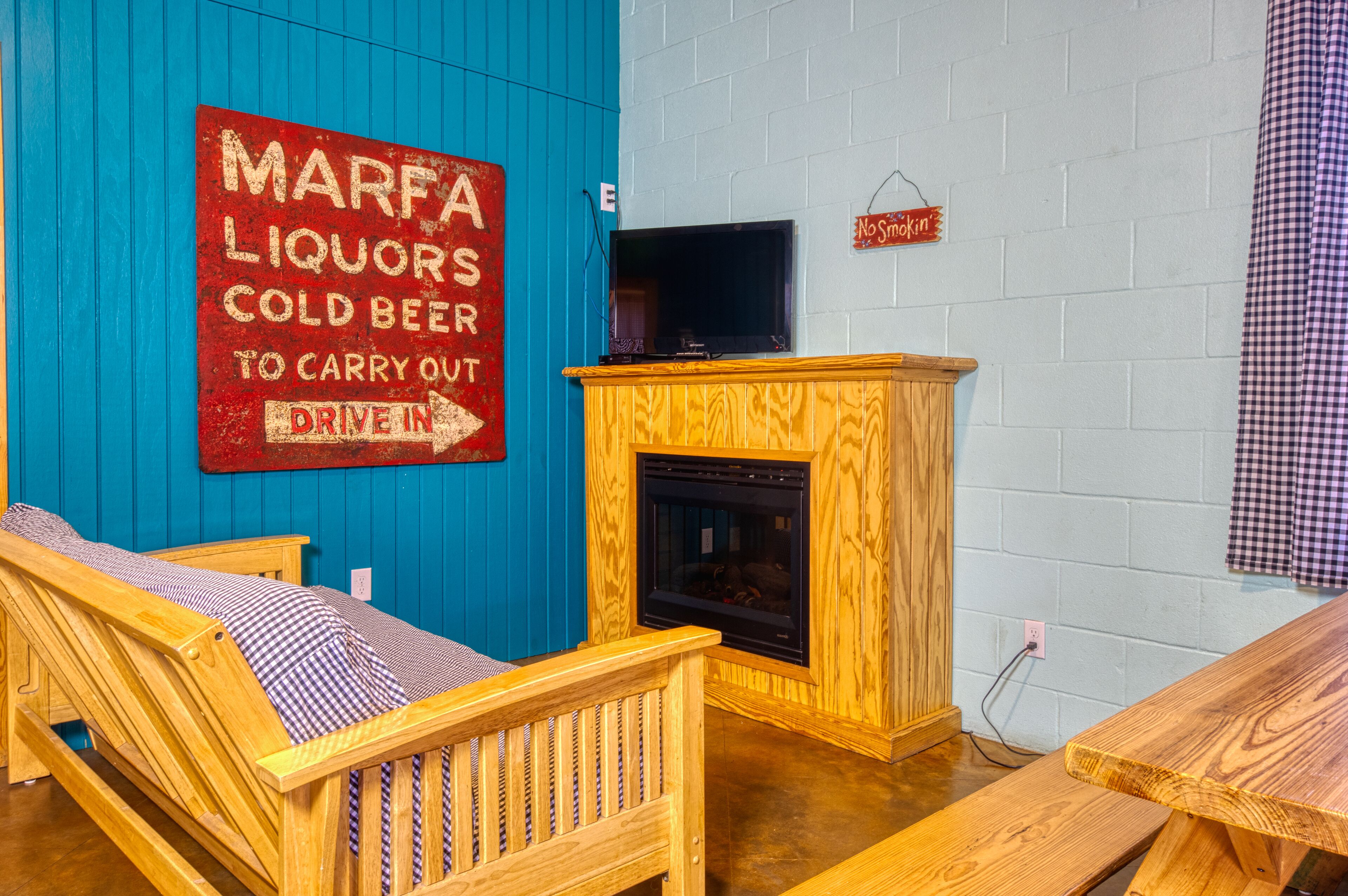 Ein Zimmer mit blauer Wand, einer Holzbank, einem Holztisch, einem Kamin und einem Schild mit der Aufschrift 'MARFA LIQUORS COLD BEER TO CARRY OUT DRIVE IN'.