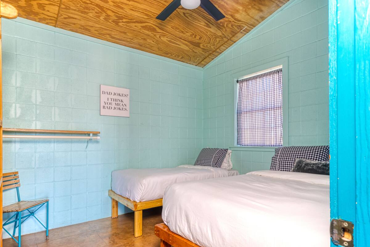 A room with two beds, a wooden ceiling, a blue wall, a window with checkered curtains, and a sign on the wall.