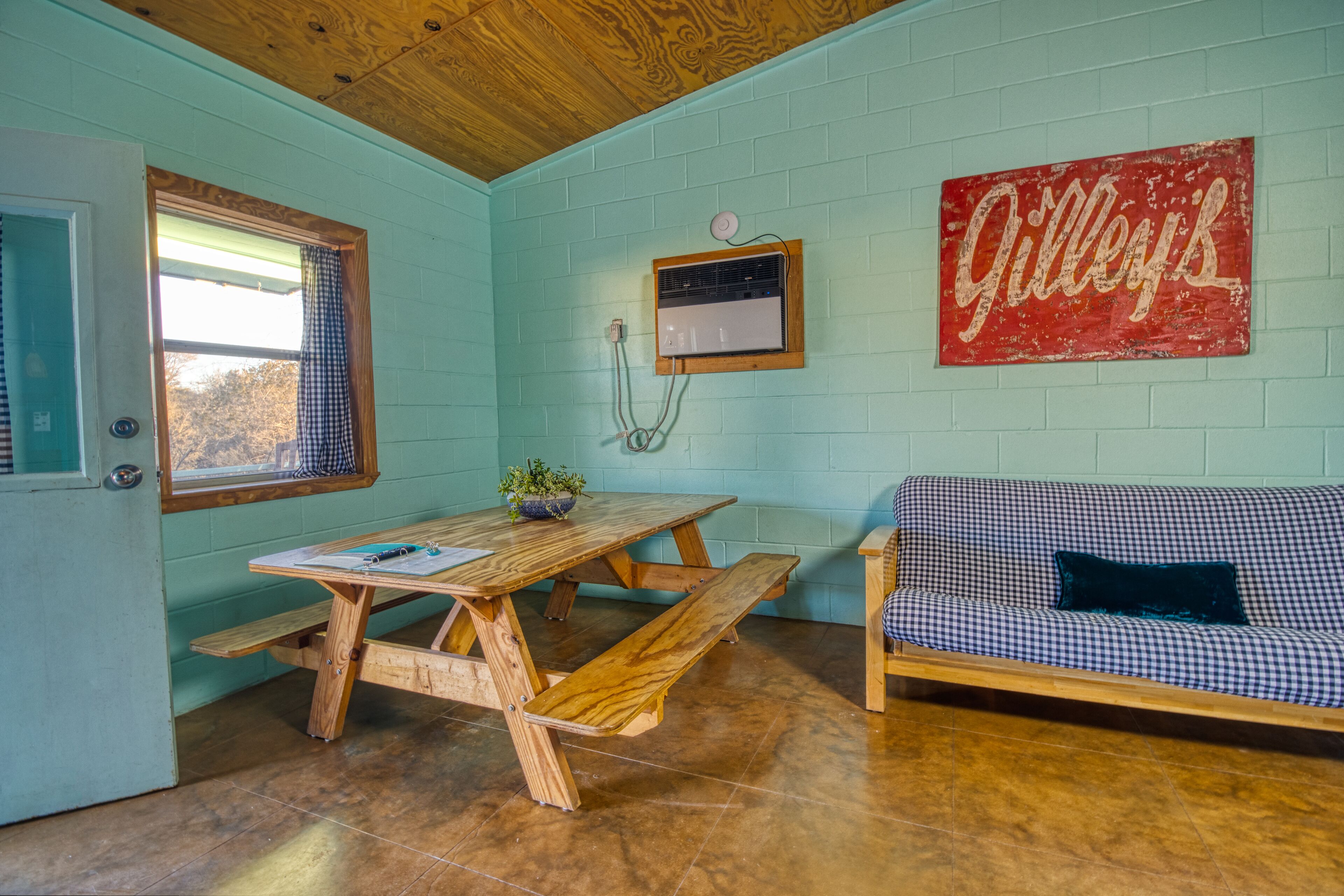 A room with a wooden table and benches, a checkered sofa, a window with curtains, and a wall-mounted air conditioning unit.