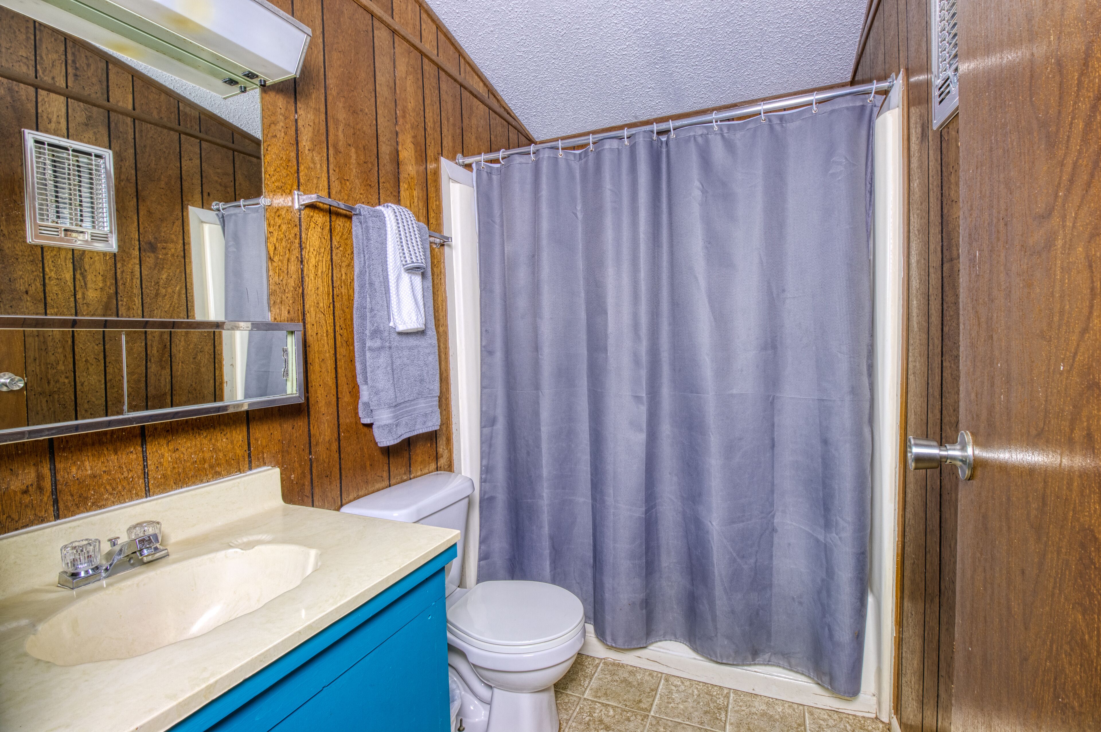 A bathroom with a wooden wall, a white toilet, a sink with a blue cabinet, and a shower curtain.