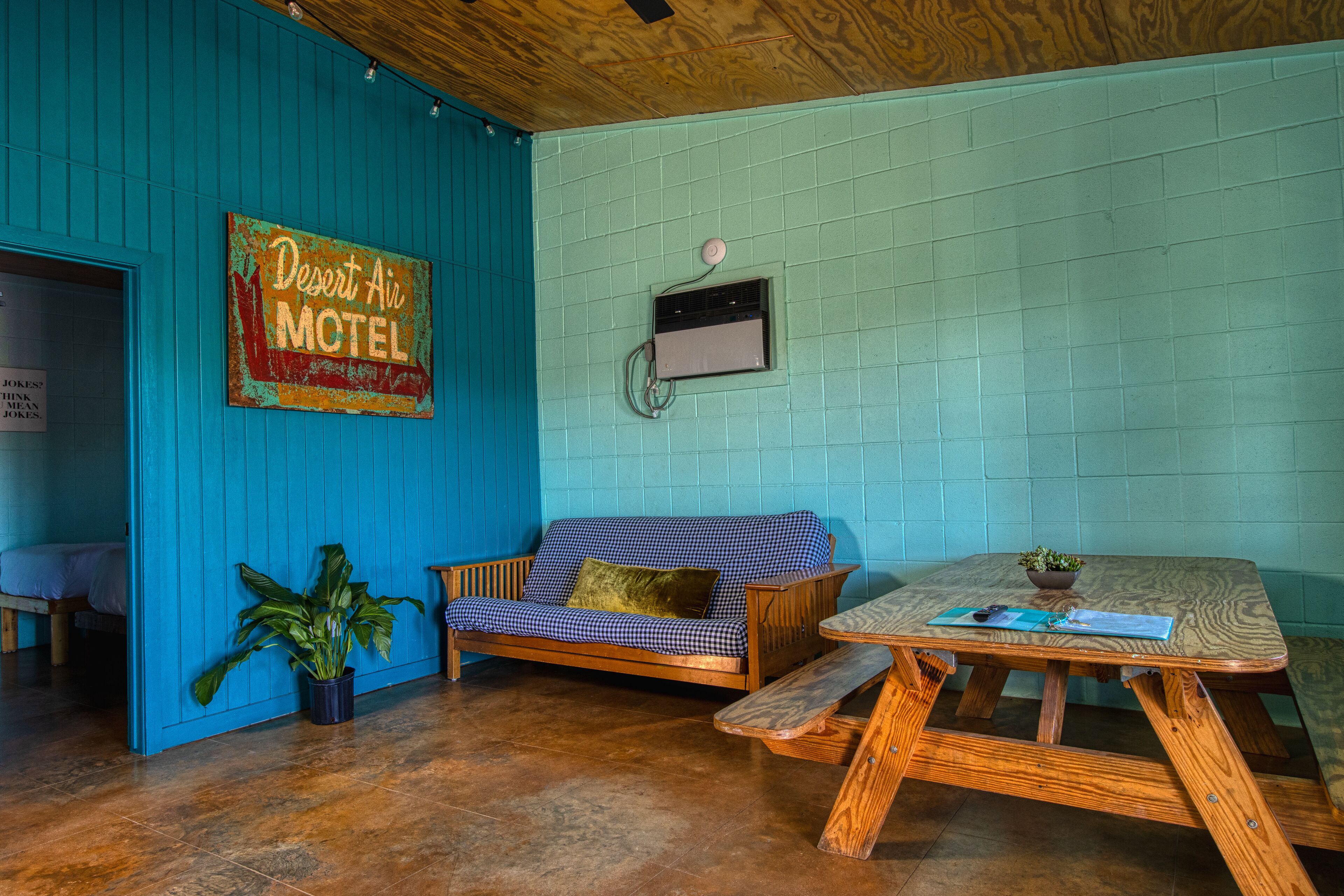 Una habitación de motel con un sofá a cuadros, una mesa de madera con un banco, una planta en maceta y un cartel en la pared.