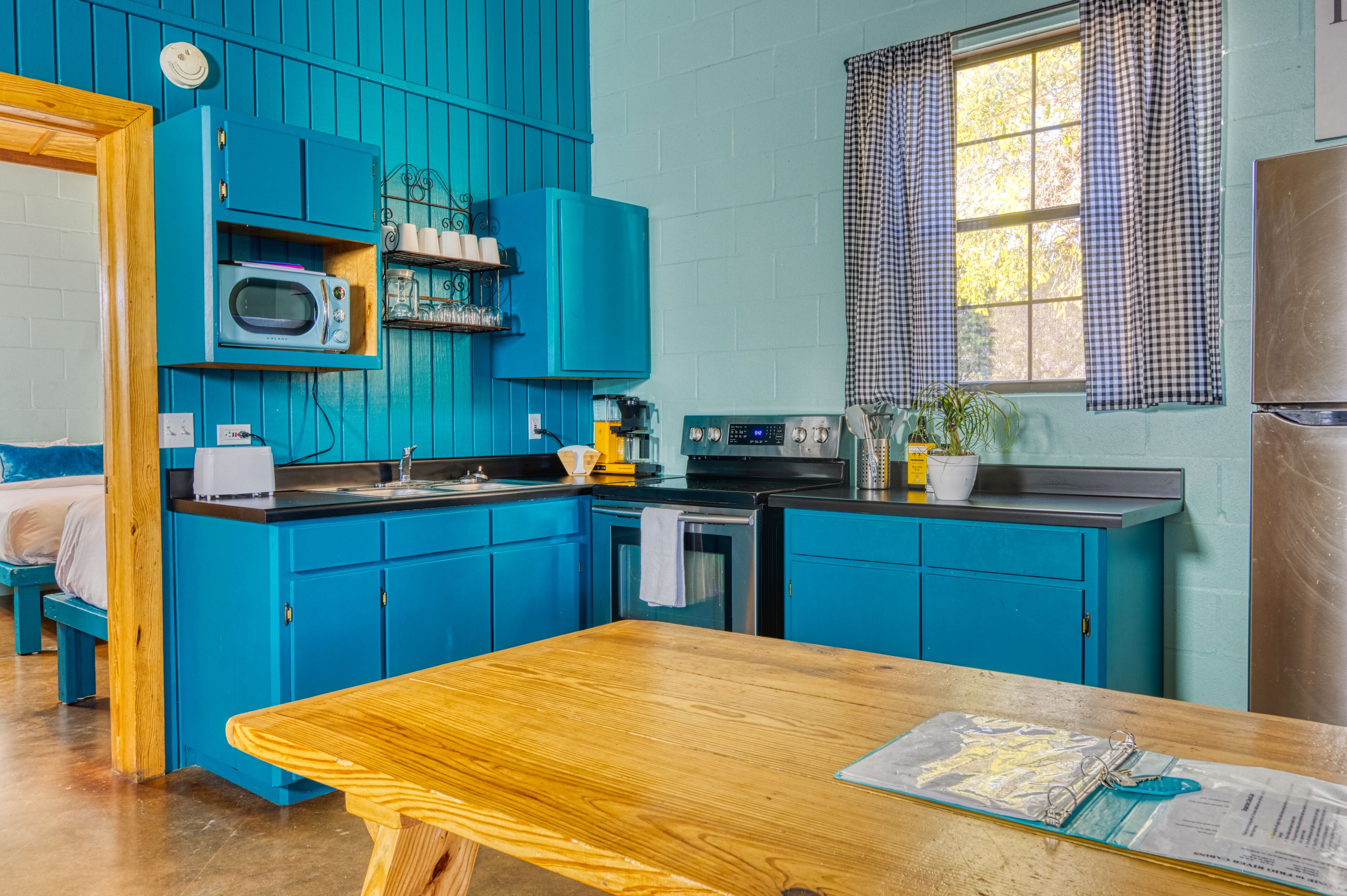 Une cuisine avec des meubles de rangement bleus, une table en bois et une fenêtre ornée de rideaux à carreaux.