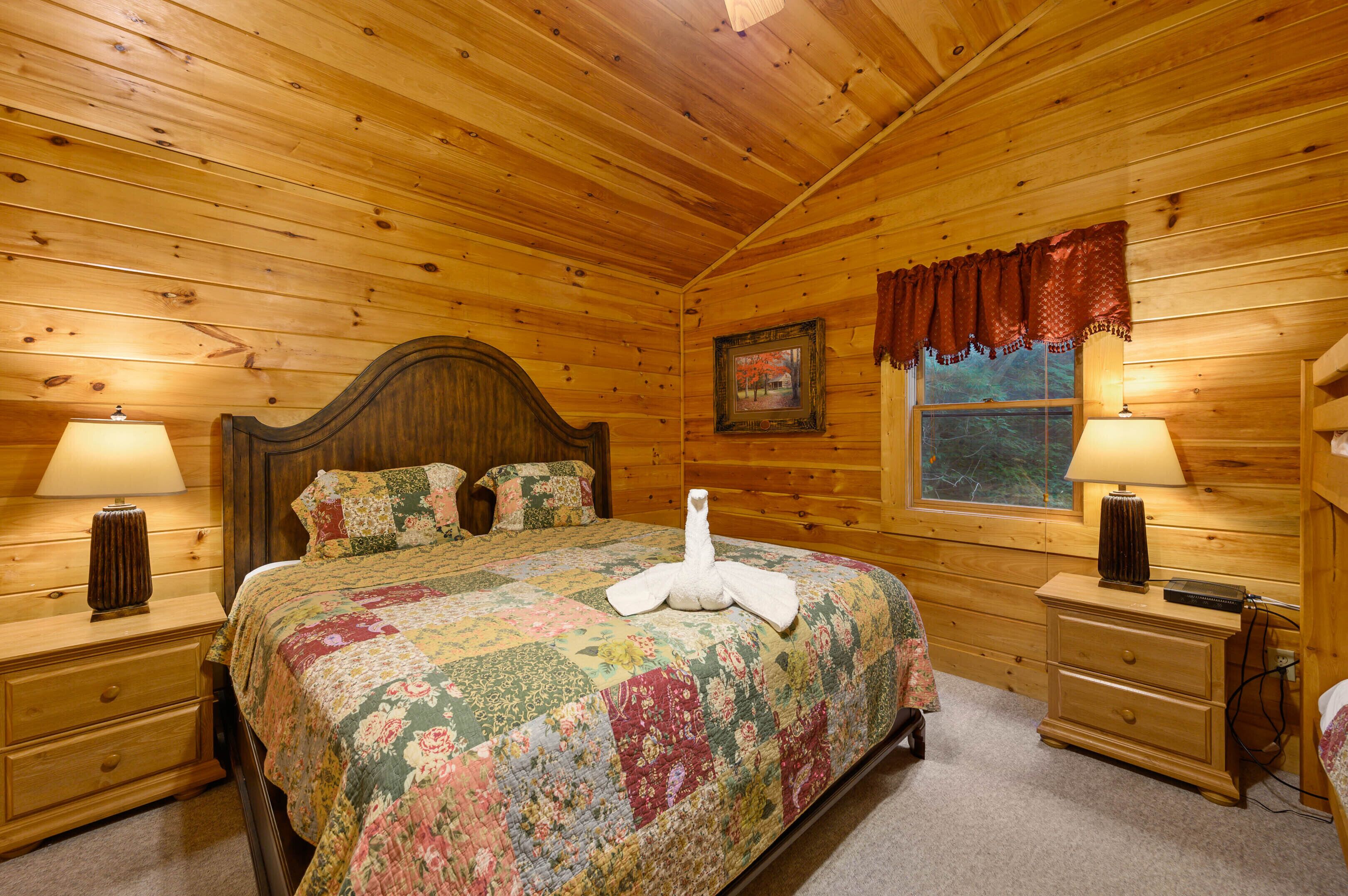 Un dormitorio de cabaña de madera con una cama grande, dos mesitas de noche con lámparas, una ventana con vistas y un cabecero de madera.