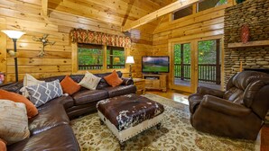 Ein Holzblockhaus-Interieur mit einer Ledersofa, einem Hocker und einem Relaxsessel, einem Fernseher und einem Kamin aus Naturstein.