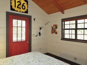 A room with a red door, a bed with a floral bedspread, a wall-mounted number sign, and a window with blinds.