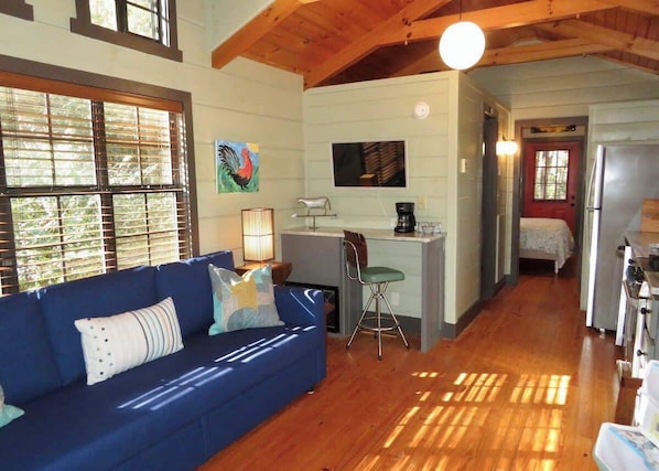 A cozy living room with a blue sofa, wooden floors, and a kitchen area with a refrigerator and a sink.