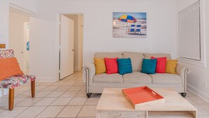 A living room with a beige sofa, colorful cushions, a wooden coffee table, a bench with a patterned cushion, and a beach-themed painting on the wall. - Beach Babe -  3/2 1st Floor Dog Friendly (Duplex) (Galveston)
