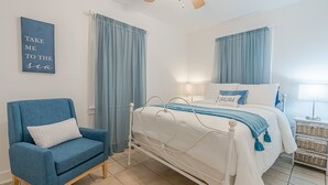A bedroom with a bed, a chair, curtains, a ceiling fan, and a wall sign. - Beach Babe -  3/2 1st Floor Dog Friendly (Duplex) (Galveston)