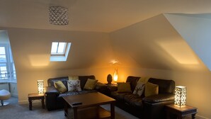 A cozy living room with a dark leather sofa, a wooden coffee table, and two side tables with lamps. A skylight provides natural light, and a fireplace adds warmth. - Ayr Loft Apartment With Fabulous Views (Ayr)