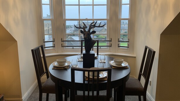 A dining area with a round table, four chairs, a deer head centerpiece, and large windows with a view of buildings and a body of water. - Ayr Loft Apartment With Fabulous Views (Ayr)