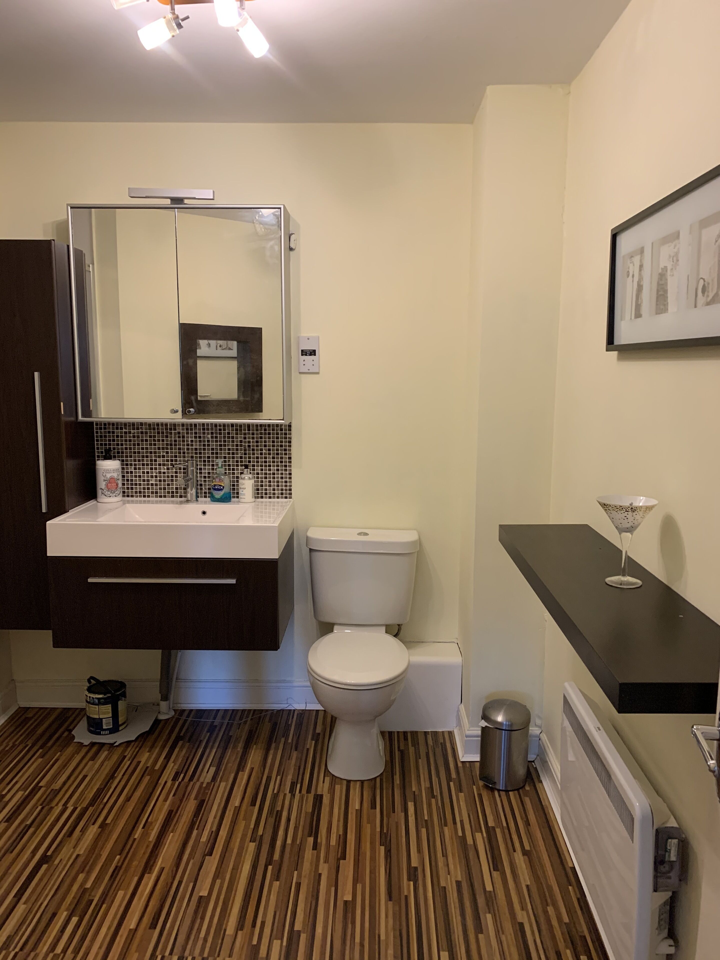 Et moderne badeværelse med trægulv, et hvidt toilet, en vask med spejl og en sort bordplade med glaspynt.