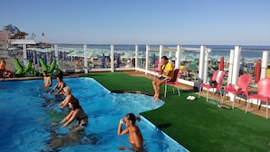 Un área de piscina con gente nadando y sentada en sillas, rodeada de sombrillas y vistas al mar.