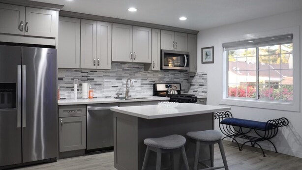 A modern kitchen with grey cabinets, a stainless steel refrigerator, a microwave, and a large island with stools.