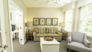 Un soggiorno con un divano color beige, un tavolino da caffè bianco e una poltrona a righe. Si vedono quadri appesi al muro e una ventola a soffitto.