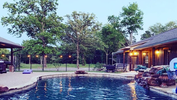 Una piscina con solárium de madera, zona de estar cubierta y un edificio con fachada rústica.
