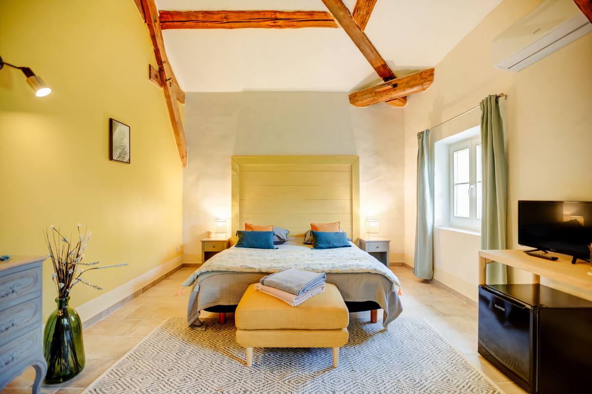 Een slaapkamer met een bed, nachtkastjes, een televisie, een dressoir, een vaas en een raam met gordijnen.