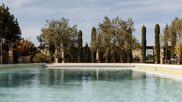 Une piscine à l’eau d’un bleu cristallin, entourée de grands arbres élancés et d’un mur de pierre.
