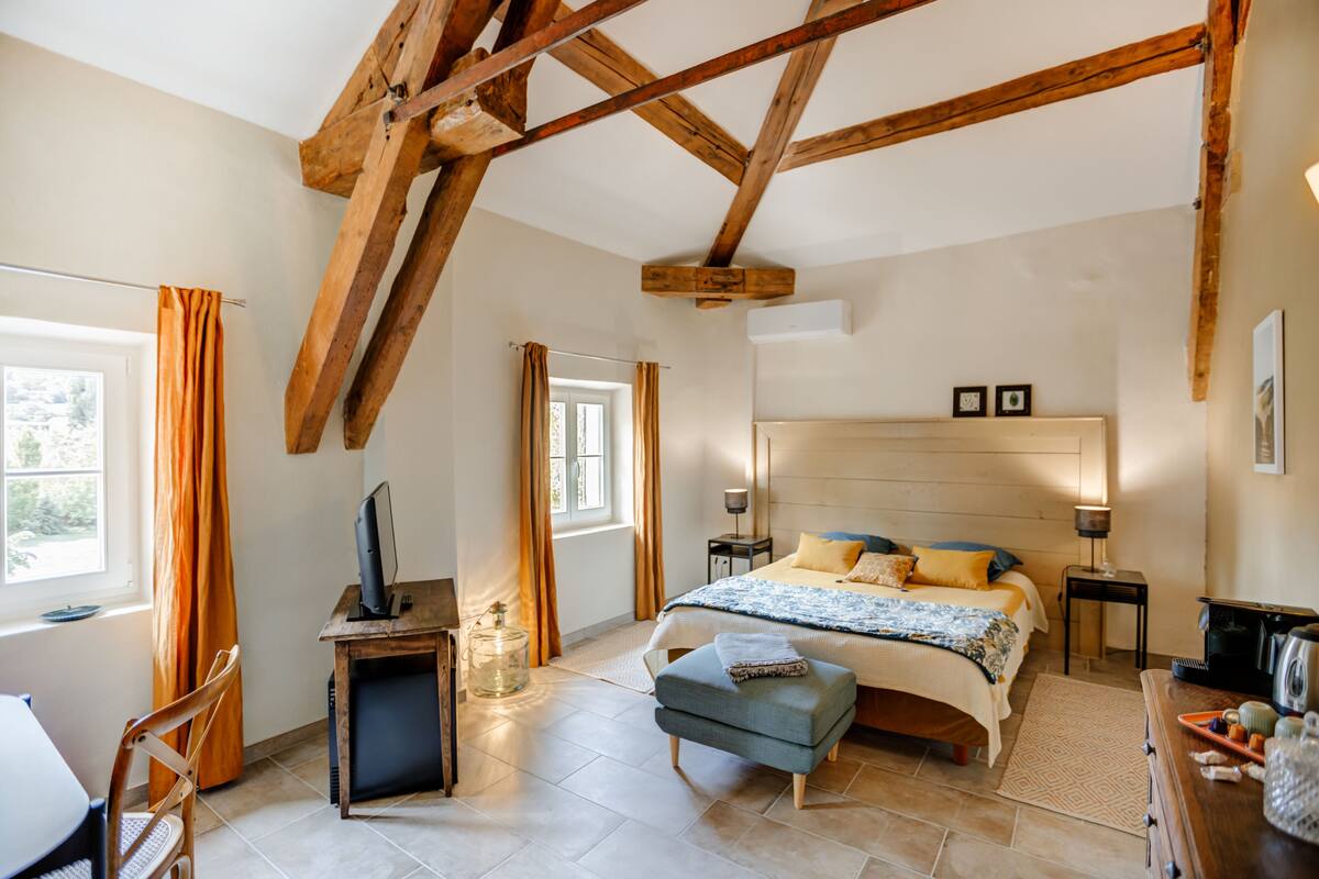 Een slaapkamer met een houten plafond, een bed met hoofdeinde, een bureau met een computer, en een raam met gordijnen.