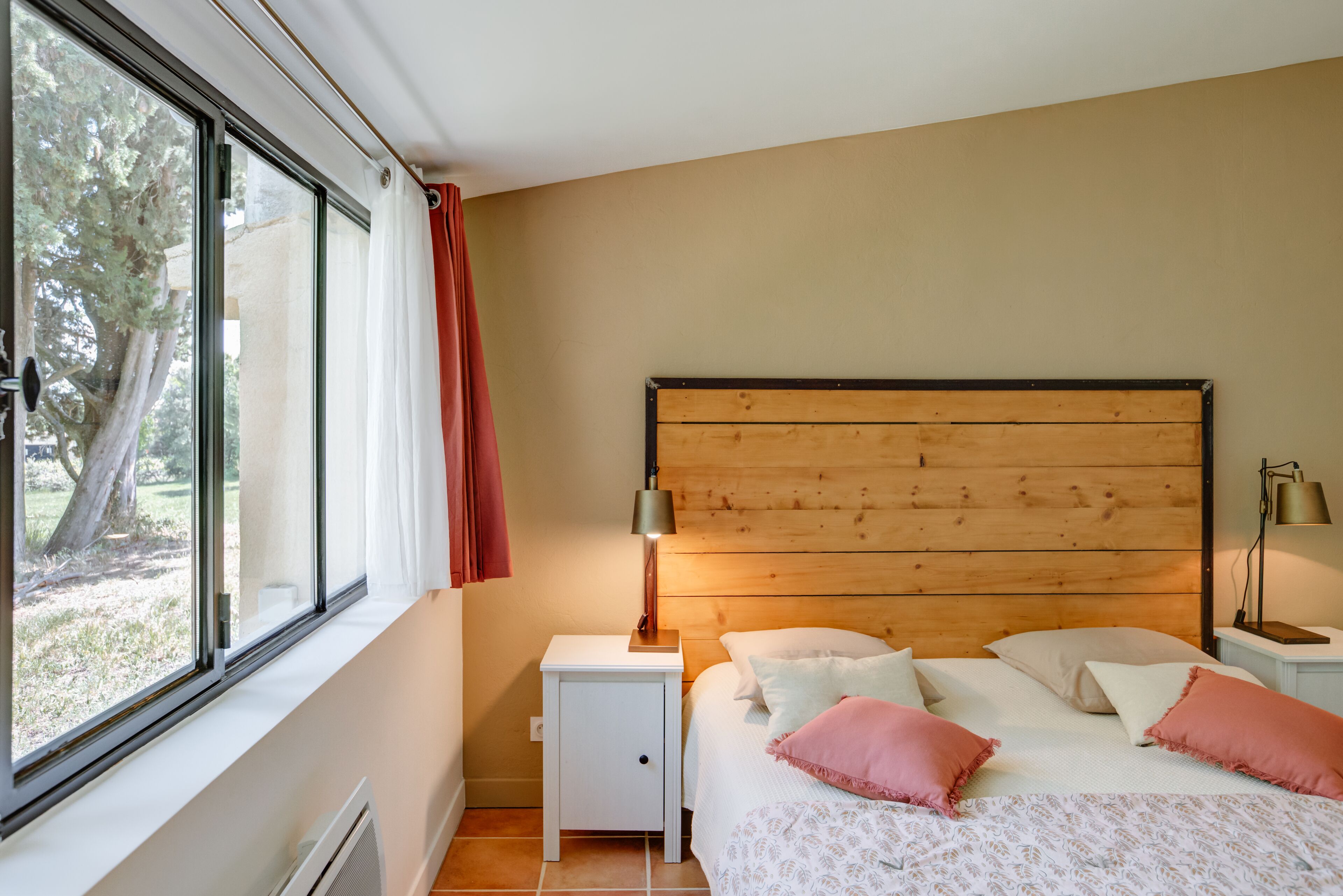 A bedroom with a wooden headboard, a white bedside table, a lamp, and a bed with patterned bedding and pink pillows.