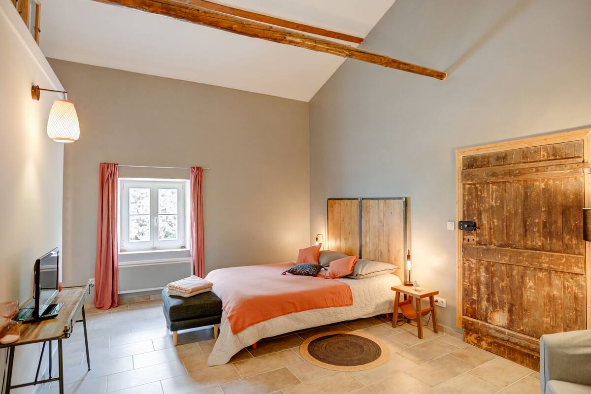 Een slaapkamer met een bed, nachtkastje, lamp, raam met gordijnen, houten deur en een klein bureau met stoel.