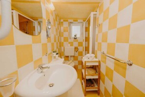 Un baño con azulejos blancos y amarillos a cuadros, un lavamanos blanco, un espejo, un inodoro y un estante pequeño con toallas.