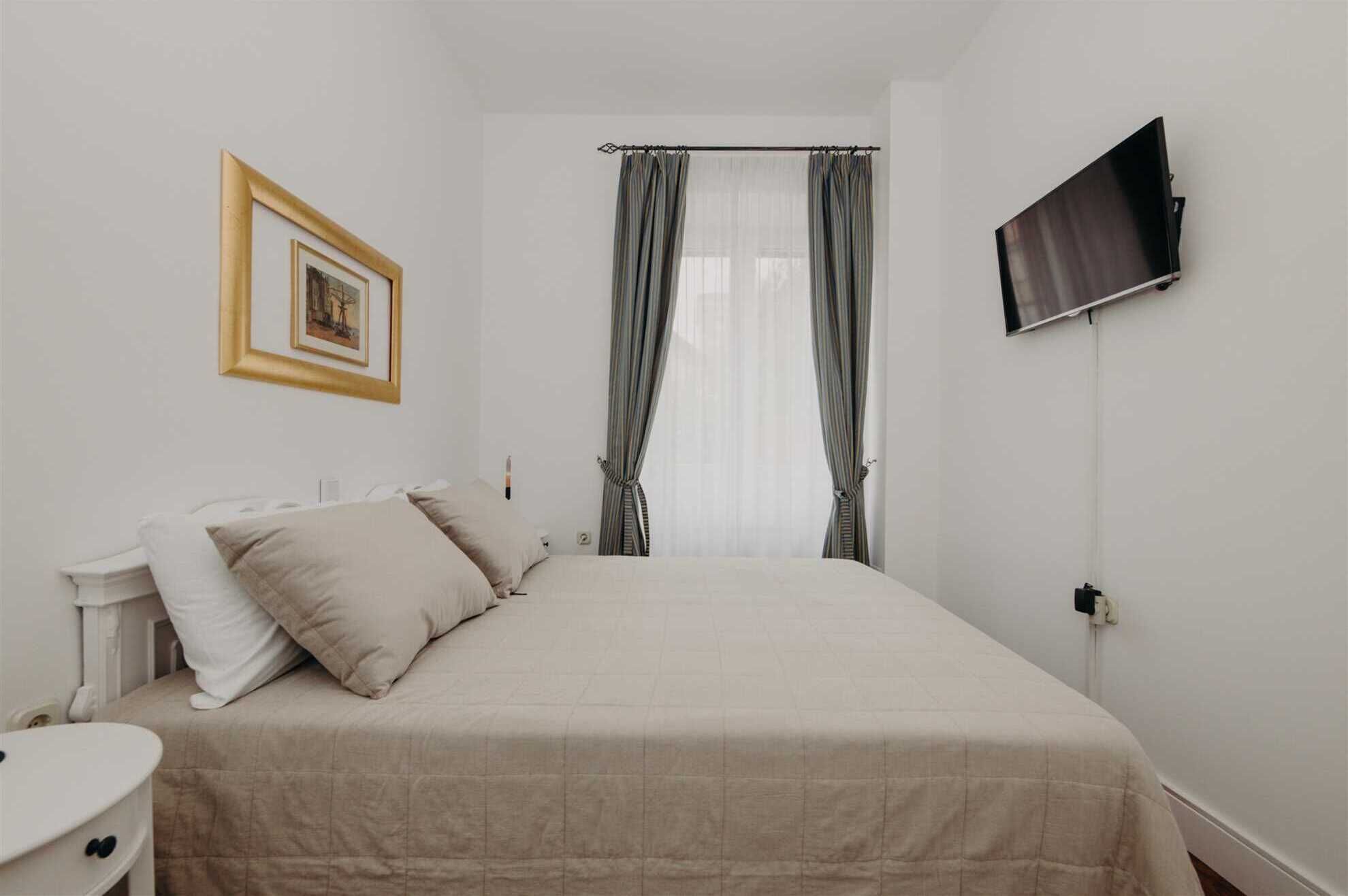 A bedroom with a bed, a bedside table, a TV mounted on the wall, and a window with curtains.