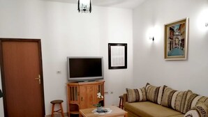 Un salon avec un canapé, un téléviseur posé sur un meuble en bois, une petite table avec une plante et un tableau encadré accroché au mur.