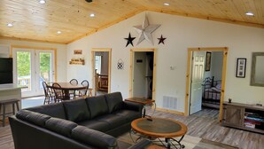 A spacious living room with a black leather sofa, a wooden coffee table, and a ceiling fan. There is a kitchen area with a dining table and chairs, and a laundry room visible through an open door.