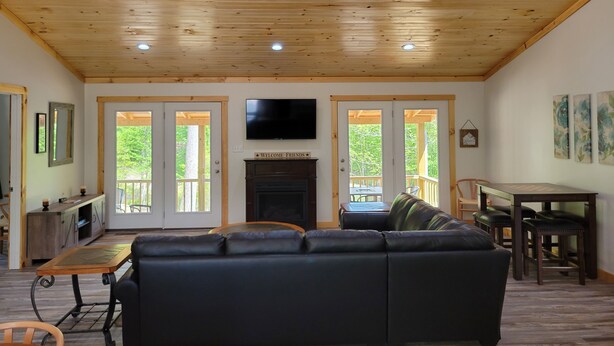 A spacious living room with a wooden ceiling, a black leather sofa, a fireplace, and a television mounted on the wall.